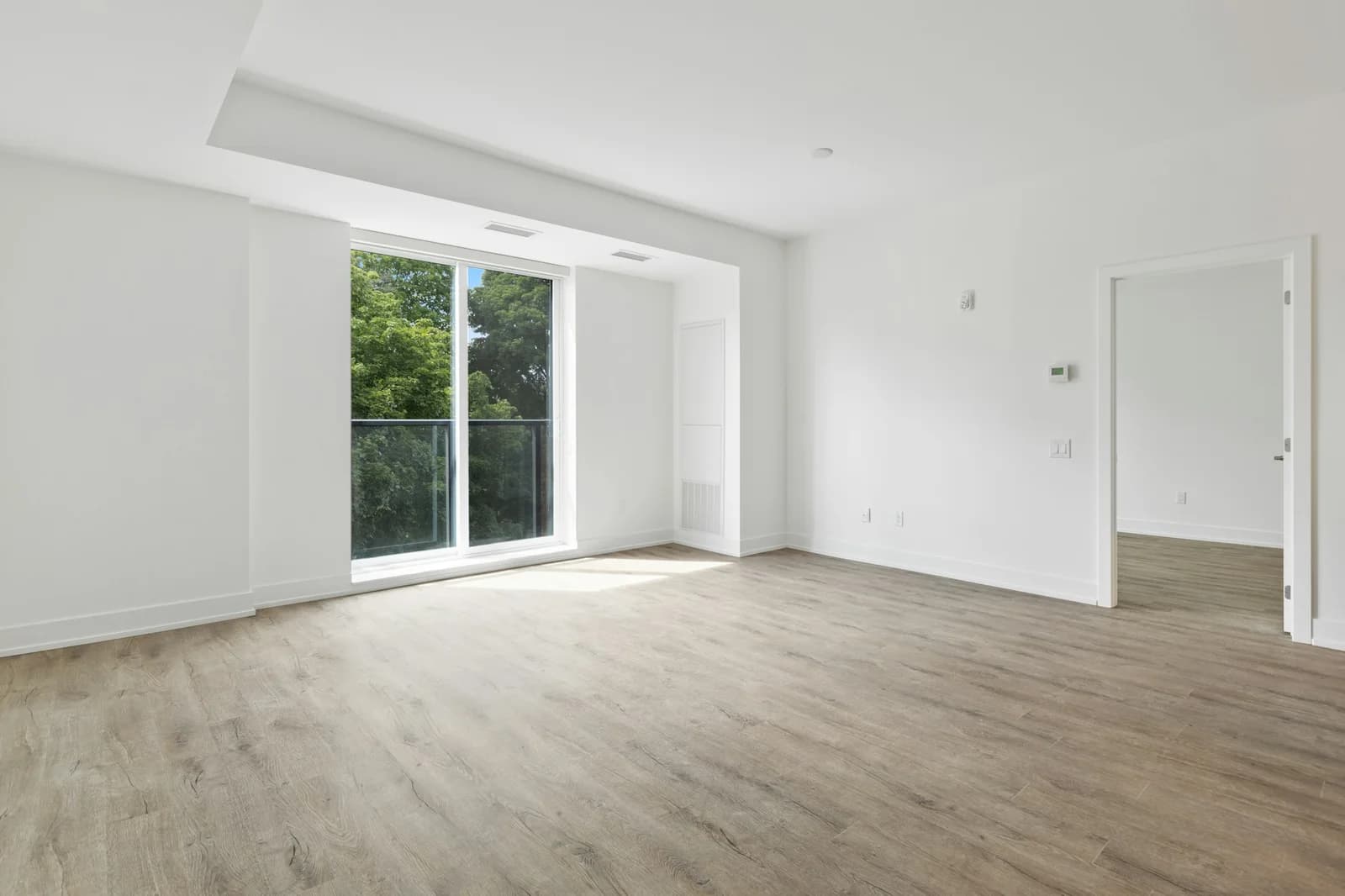 Séjour vide d’un appartement neuf aux murs blancs et au parquet clair, éclairé par une grande baie vitrée donnant sur des arbres.