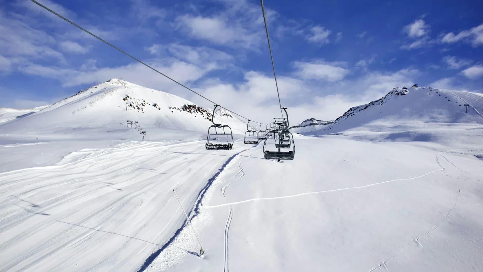 Télésiège vide qui s’élève au-dessus d’une piste de ski damée, entre des sommets enneigés, sous un ciel bleu parsemé de nuages blancs.