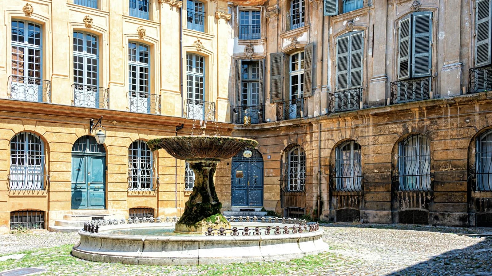 Fontaine de pierre au centre d’une place pavée du vieil Aix-en-Provence, entourée de façades ocre aux volets bleus, sous une lumière de jour