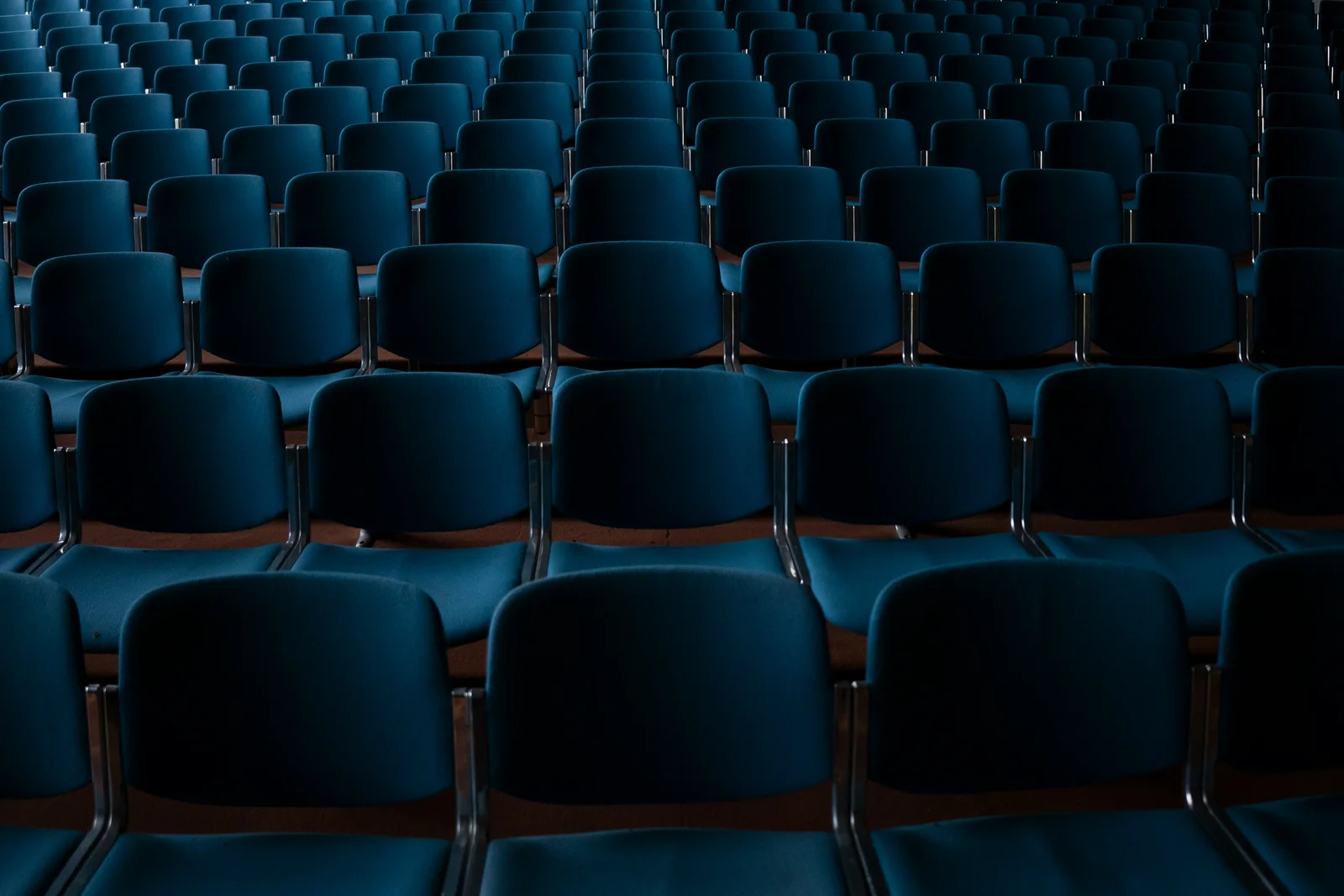 Des rangées de chaises bleues vides, alignées dans une grande salle de conférence peu éclairée.