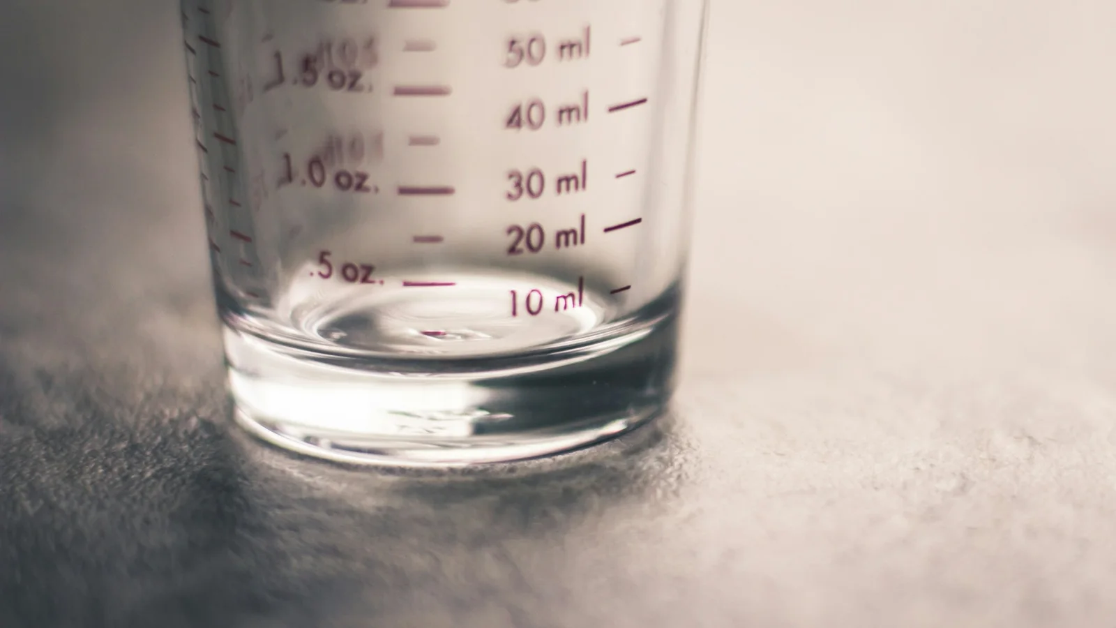Un verre doseur en verre, gradué en millilitres et en onces, posé vide sur une surface grise, sous une lumière douce.
