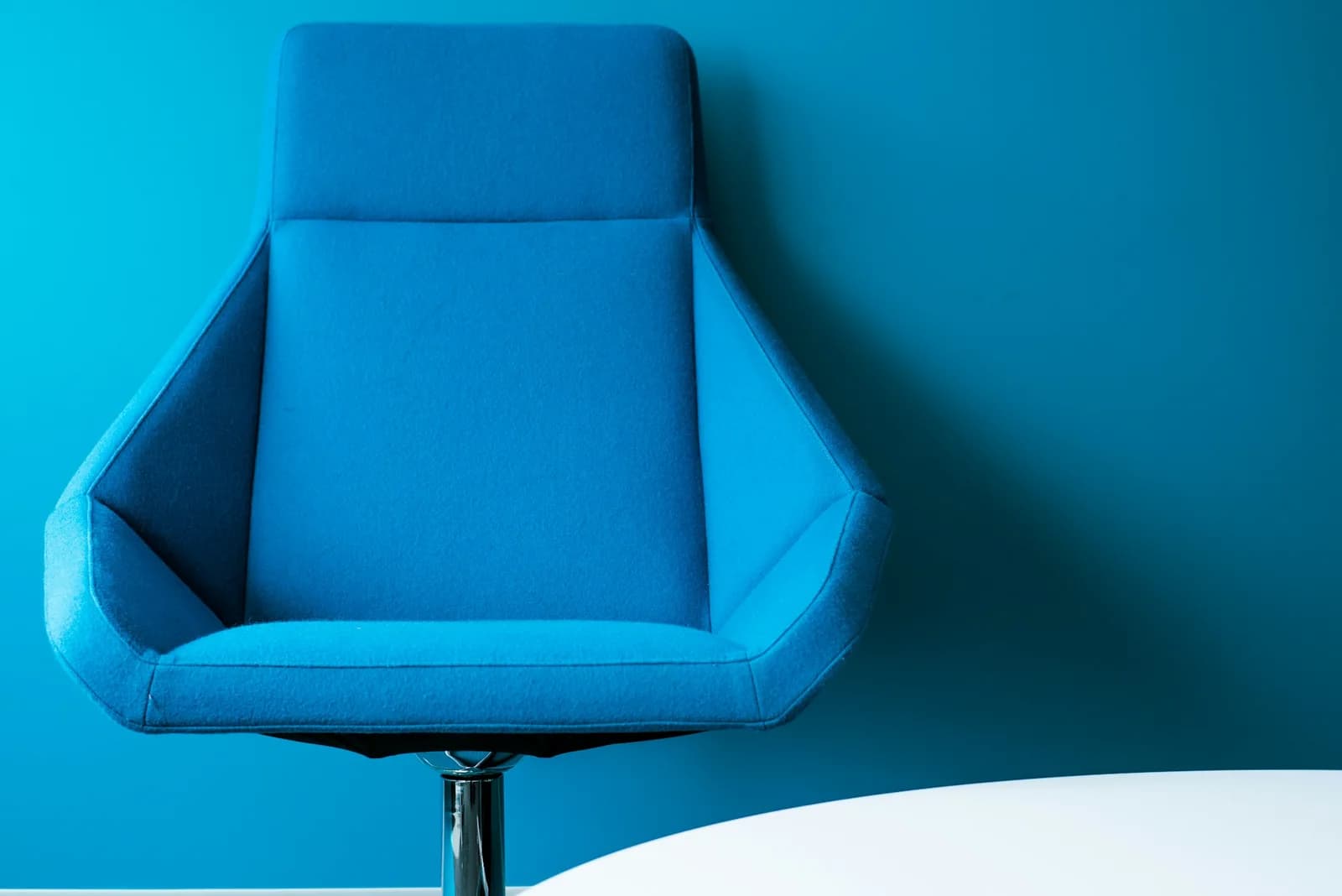 Fauteuil pivotant bleu vide devant un mur bleu, avec le bord d’une table blanche au premier plan