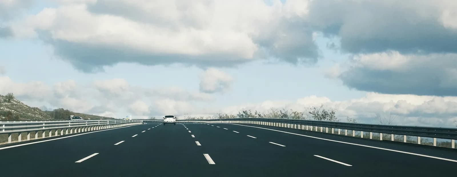 Une autoroute presque déserte sous un ciel bleu chargé de nuages blancs, une voiture blanche au loin