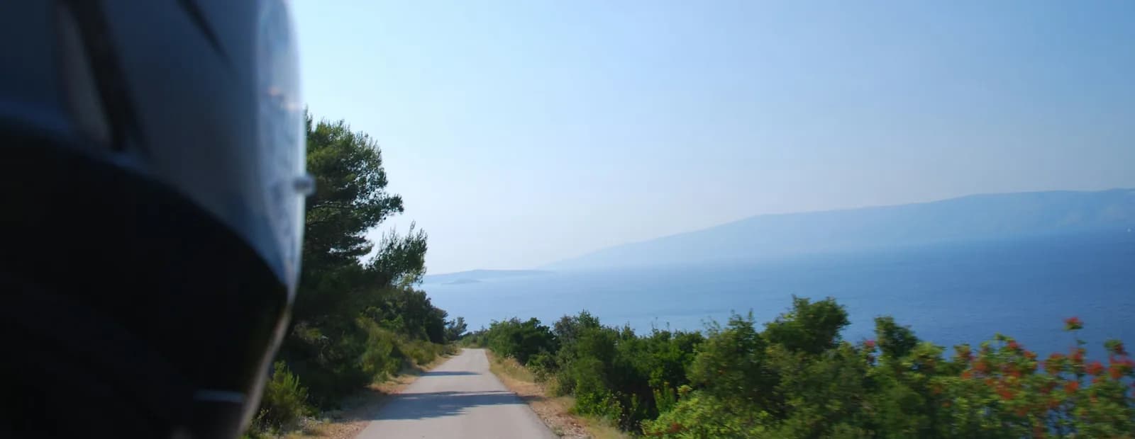 Route côtière étroite vue depuis un deux-roues, le casque du conducteur au premier plan, face à une mer bleue et à une île au loin.
