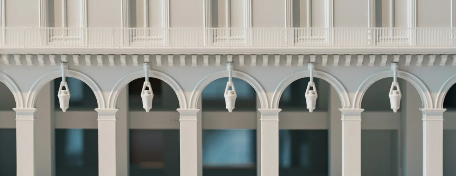 Maquette d’architecture blanche d’un immeuble à arcades, avec une galerie voûtée au rez-de-chaussée, des lanternes suspendues et une rangée de fenêtres à l’étage