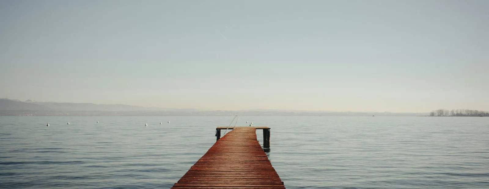 Ponton de bois qui s’avance sur un lac calme, sous un ciel pâle, la rive estompée à l’horizon.