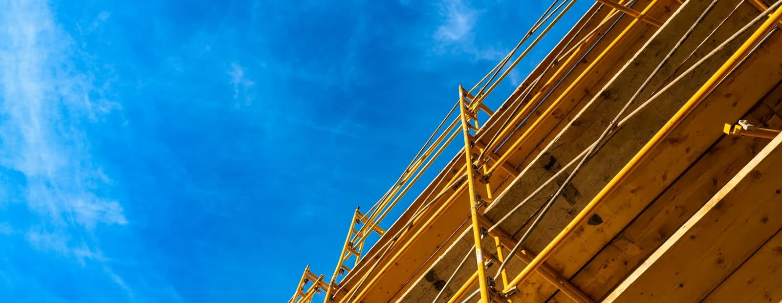 Échafaudage jaune et planches de bois d’un chantier de rénovation vus en contre-plongée sous un ciel bleu