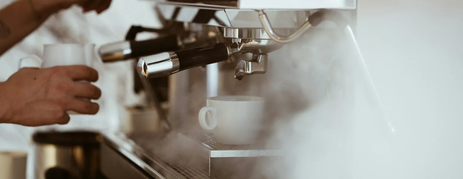 Les mains d’un barista tiennent un pichet et un porte-filtre devant une machine à expresso en inox qui fume, dans un café.