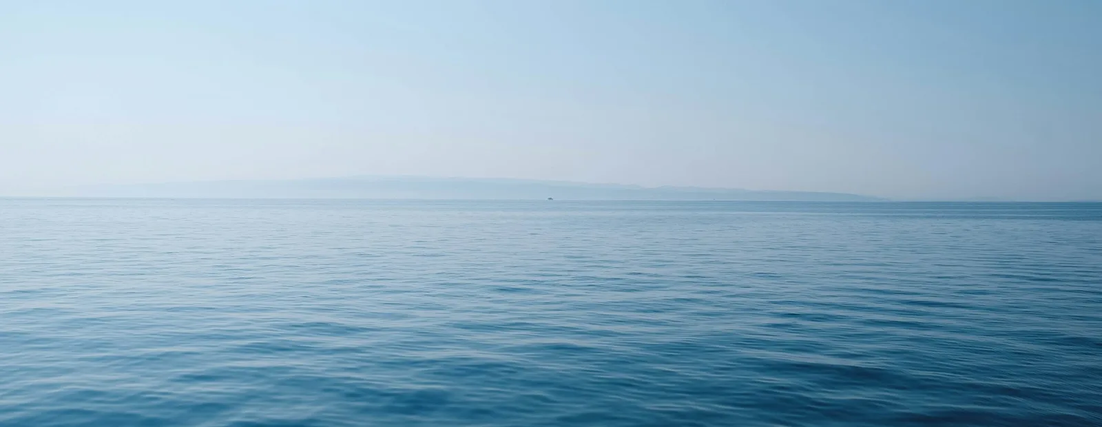 Mer calme d’un bleu profond sous un ciel pâle, avec une côte à peine visible à l’horizon.