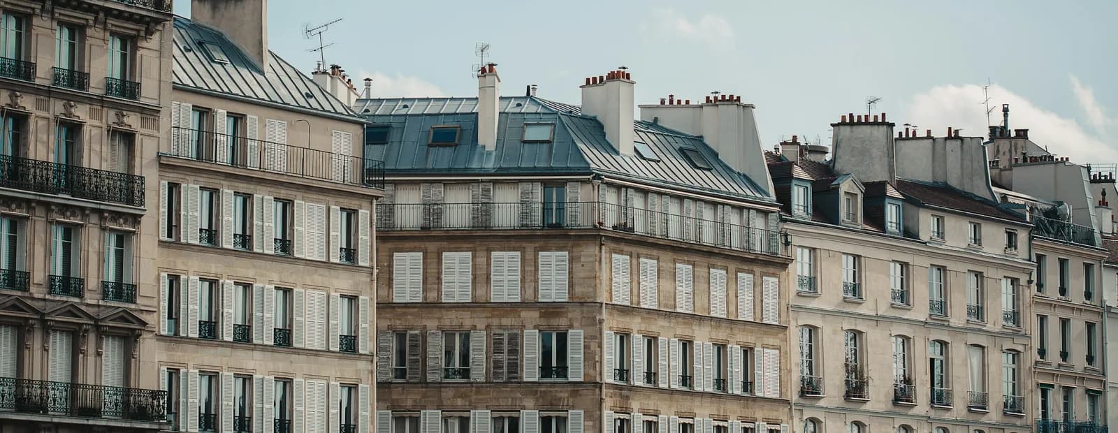 Rangée d’immeubles haussmanniens à Paris, façades claires à balcons filants et toits en zinc sous un ciel bleu voilé