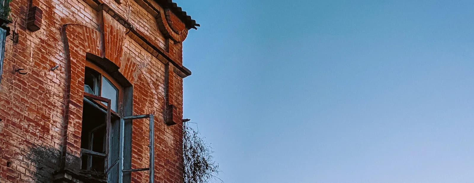 Angle d’un immeuble ancien en brique à corniche moulurée, fenêtre cintrée entrouverte, sous un ciel bleu dégagé
