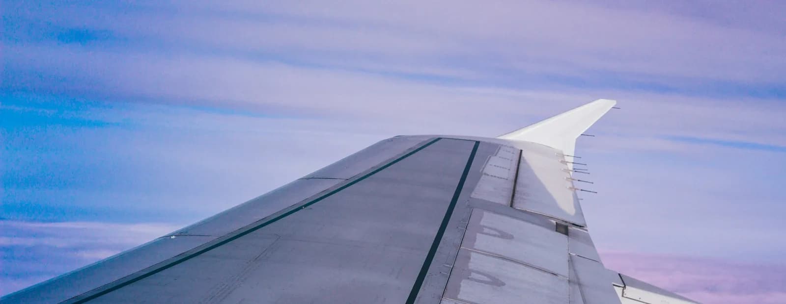 Aile d’avion grise vue depuis le hublot, avec son winglet blanc, sur un ciel voilé de nuages bleus et mauves