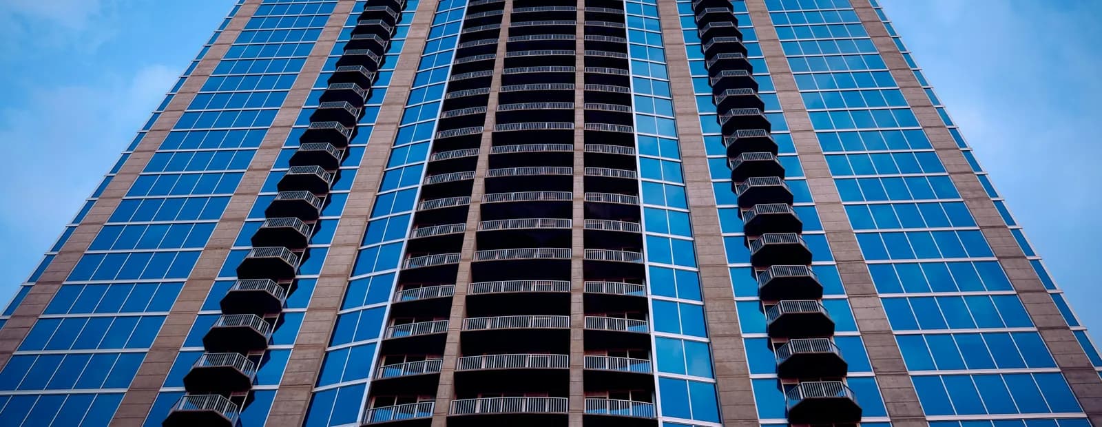 Tour d’habitation vue en contre-plongée, façades vitrées bleues et balcons superposés sous un ciel bleu nuageux