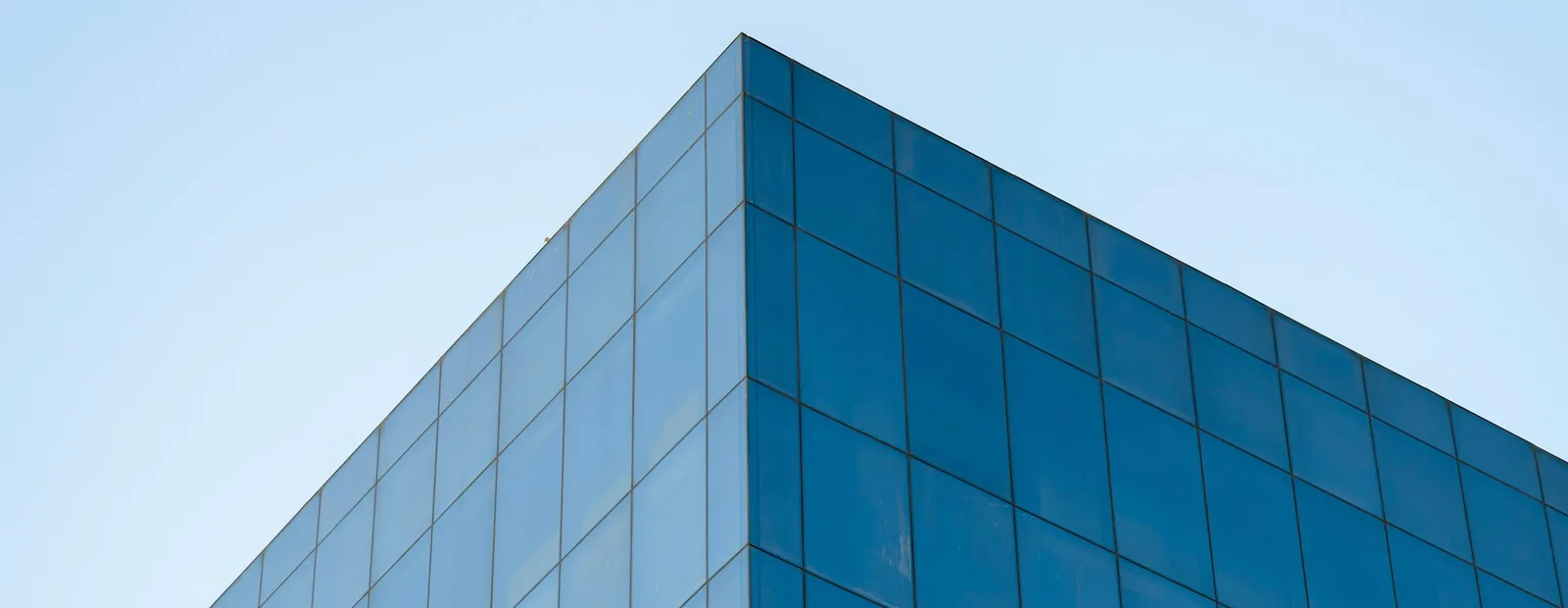 Angle d’un immeuble de bureaux entièrement vitré de panneaux bleus réguliers, vu en contre-plongée sur un ciel bleu très clair.